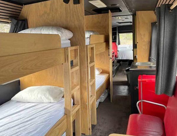 Interior view of a converted bus with two sets of wooden bunk beds on one side and a red-seated dining booth on the opposite side. A kitchenette with a red fridge and stainless steel sink is positioned beside the seating area. One of the Cabins, Dwellings, Caravans, & Glamping Tents at Stawell Grampians Gate C&C Park.
