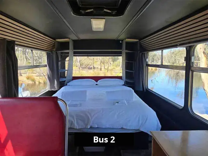 Queen bed setup at the rear of Bus 2 with panoramic windows overlooking the lake and surrounding bushland.