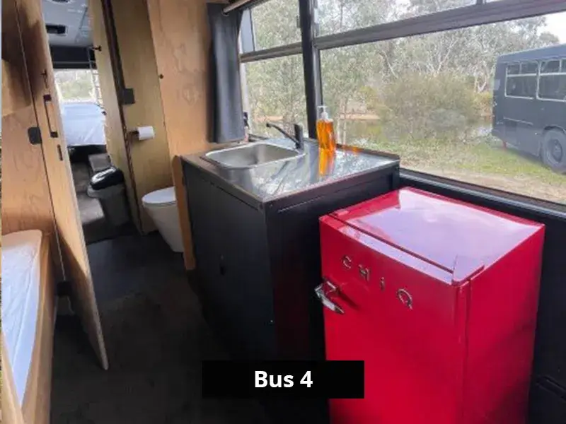 Compact kitchenette in Bus 4 featuring a stainless steel sink, bright red mini fridge, and large window with bush views.