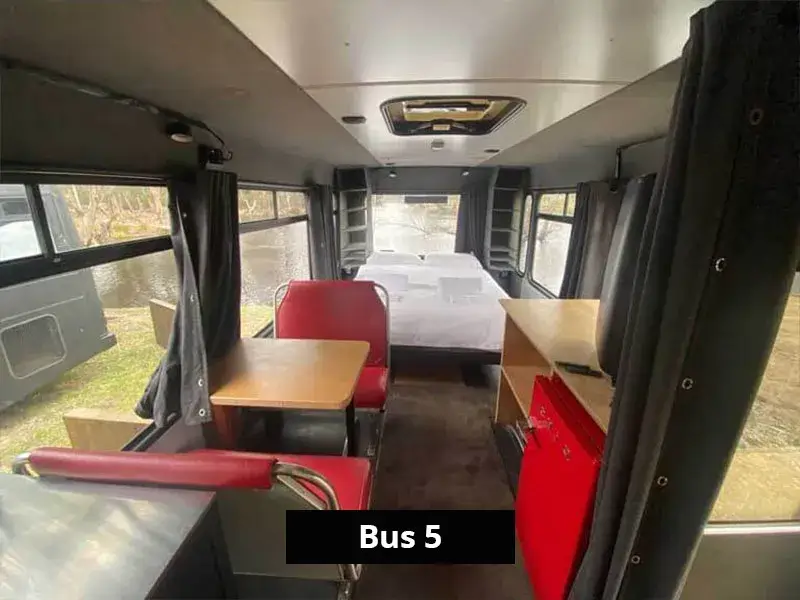 Spacious bus interior with red dinette, modern kitchenette, and queen bed overlooking lake views through rear windows.