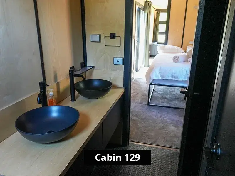 Modern bathroom in Cabin 129 with dual matte black vessel sinks, minimalist fixtures, and a clear view into the bedroom with a queen bed and natural light, located at Grampians Gate caravan park in Stawell.