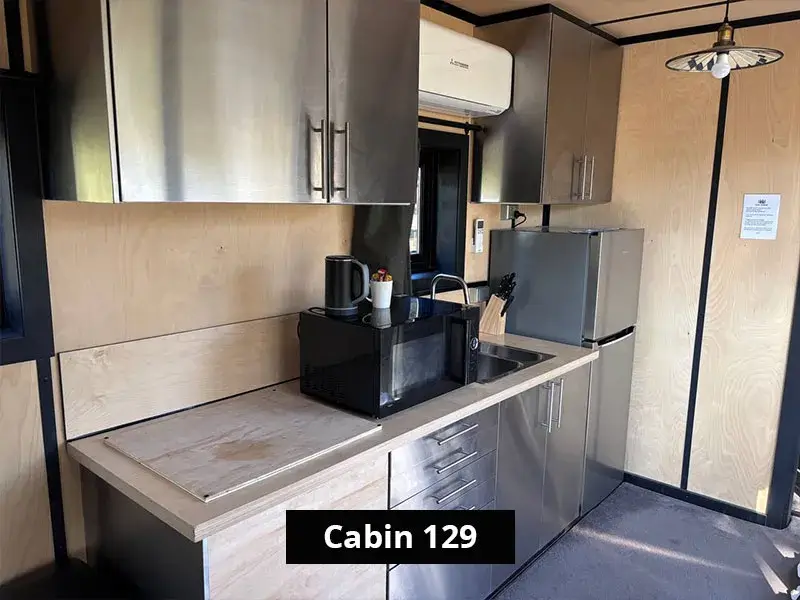Kitchenette in Cabin 129 at Stawell C&C Park Grampians Gate, equipped with stainless steel cabinetry, a microwave, electric kettle, knife set, sink, refrigerator, and overhead storage – designed for short-stay convenience.