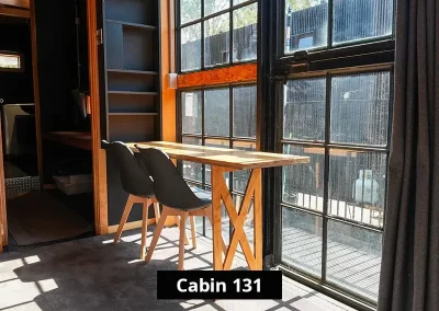 Dining or workspace area in Cabin 131 at Stawell C&C Park, with a compact wooden table and two modern chairs placed by large industrial-style windows for natural light and scenic views.