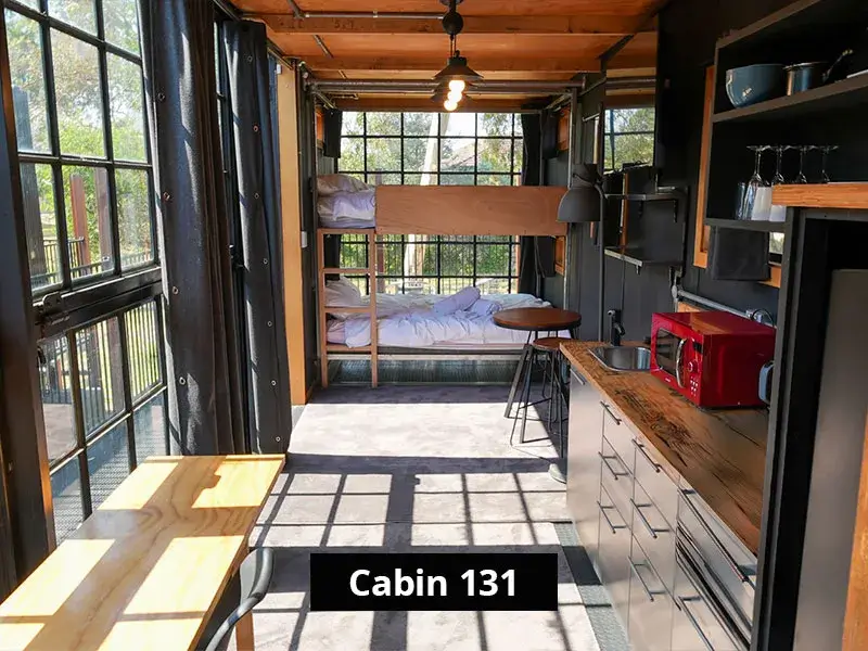 Interior of Cabin 131 at Stawell C&C Park Grampians Gate, featuring a compact layout with a timber bunk bed, modern kitchenette with a red microwave, stainless steel cabinets, open shelving, and a floor-to-ceiling window providing natural light.