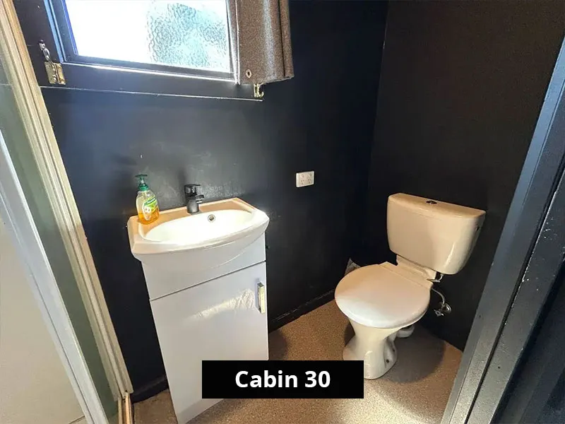 Compact bathroom in Cabin 30 at Stawell C&C Park featuring a modern white toilet and matching vanity with built-in sink, chrome tapware, hand soap, and toilet paper holder against a dark wall.