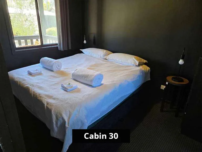 Queen bedroom in Cabin 30 with a large window, soft lighting, dark walls, a queen-size bed dressed in crisp white linens and towels, and dual bedside tables with hanging lamps.