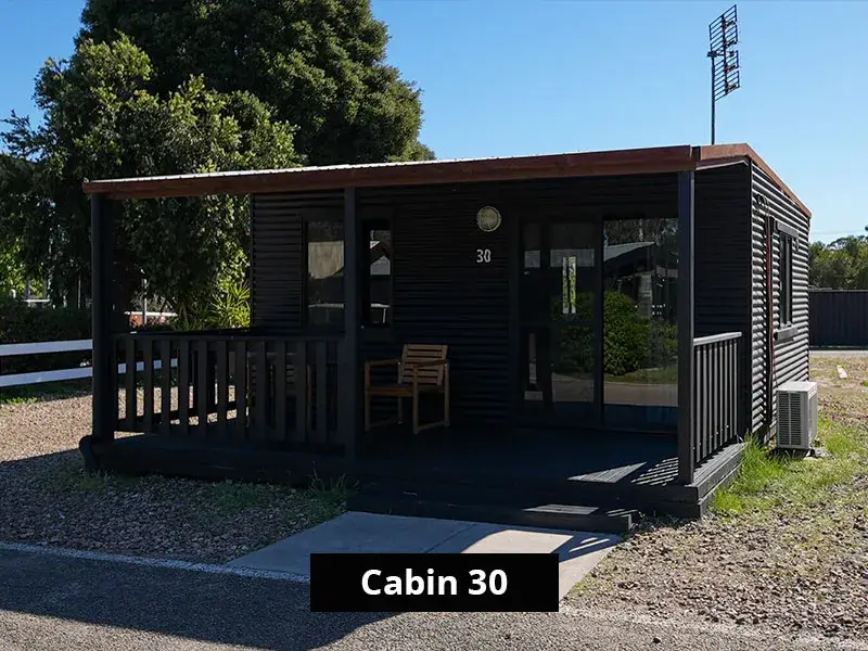 Exterior view of Cabin 30 with private deck and outdoor seating at Grampians Gate C&C Park. Set in a peaceful location, this compact black-painted cabin features a shaded porch, modern lines, and nearby nature—ideal for travelers seeking a simple, self-contained stay.