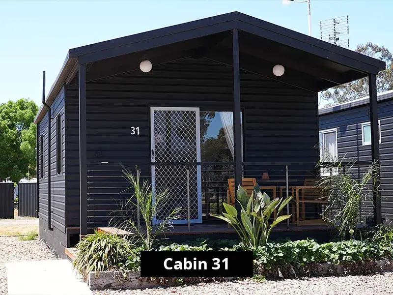 Front view of Cabin 31 at Stawell C&C Park, featuring a black weatherboard exterior, covered veranda with seating, and surrounding greenery.