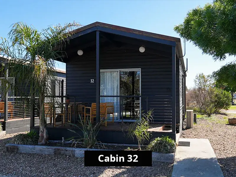 Front view of Cabin 32 at Stawell C&C Park, with dark timber siding, sliding glass door, private deck and outdoor seating surrounded by native landscaping.