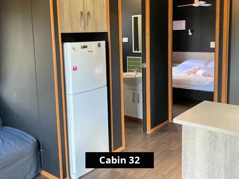 A look inside Cabin 32 featuring a full-size fridge, modern cabinetry, and access to the bathroom and main bedroom, with dark feature walls and timber accents.
