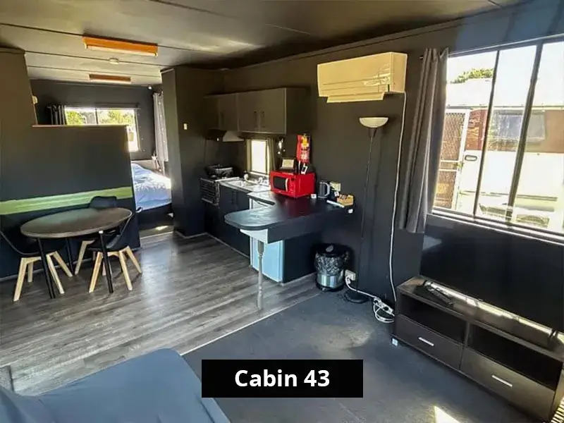 Interior of Cabin 43 displaying an open-plan layout with kitchenette, dining area, air conditioning, microwave, TV unit, and natural light through large windows—perfect for short-term or extended stays.