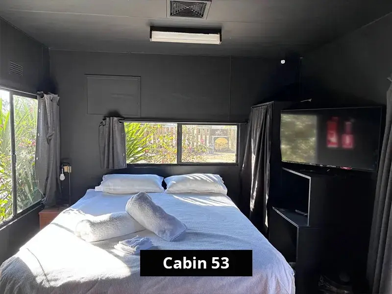 Queen bedroom inside Cabin 53 at Stawell C&C Park, with dark walls, wide windows, a mounted flat-screen TV, and a queen bed made with white linen and rolled towels.