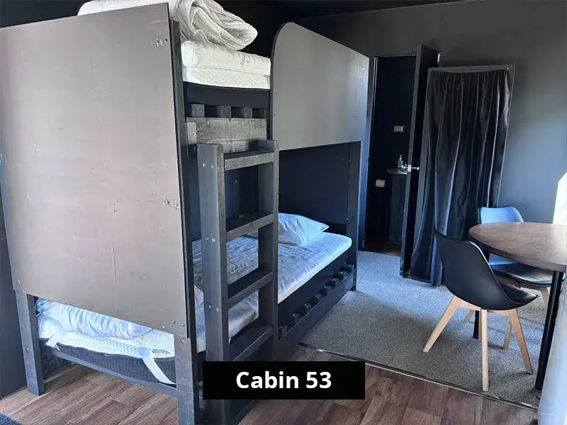 Bunk beds and dining area inside Cabin 53, featuring dark timber bunk beds, round dining table with chairs, curtain-divided bathroom access, and compact interior layout ideal for group stays.