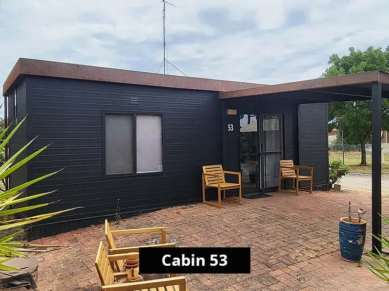 Exterior of Cabin 53 at Stawell C&C Park Grampians Gate, showing a compact dark timber-clad cabin with a small brick patio, wooden outdoor seating, and a flat roof design surrounded by landscaped greenery.