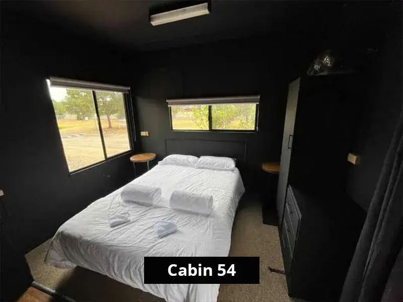 Queen bedroom in Cabin 54 with dark painted walls, large windows on two sides, a queen bed dressed in white linen and towels, and two small round side tables.