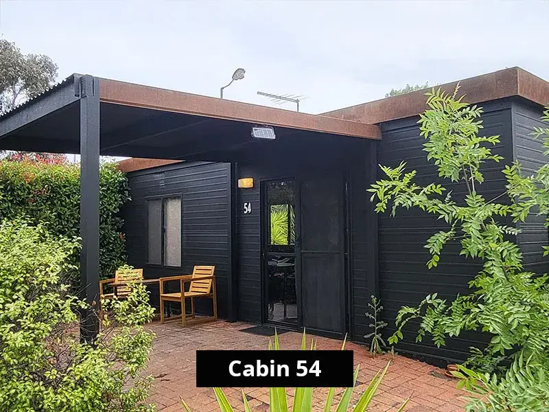 Exterior of Cabin 54 at Stawell C&C Park Grampians Gate, showing a modern black timber cabin with outdoor seating on a paved patio, surrounded by garden plants and a covered entrance.