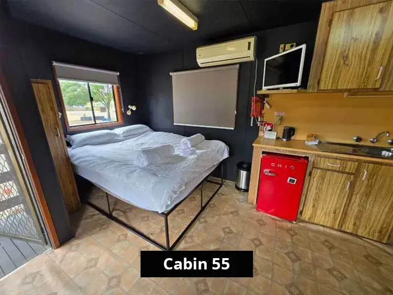 Interior of Cabin 55 at Stawell C&C Park featuring a queen bed with white linen, air conditioning, wall-mounted TV, timber-style kitchenette with red mini fridge, microwave, sink, and natural light from two windows.