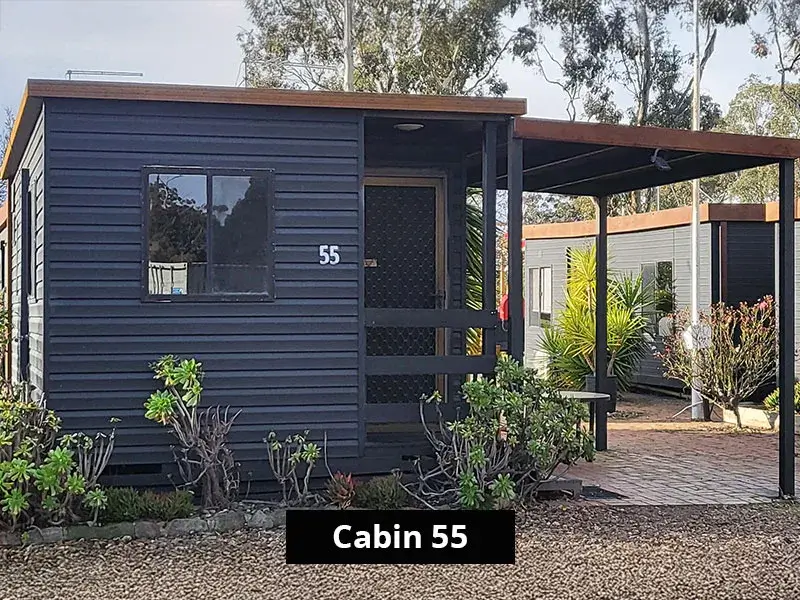 Exterior of Cabin 55 at Stawell C&C Park, featuring a dark timber cabin with a covered porch, garden landscaping, and cabin number visible on the front door.
