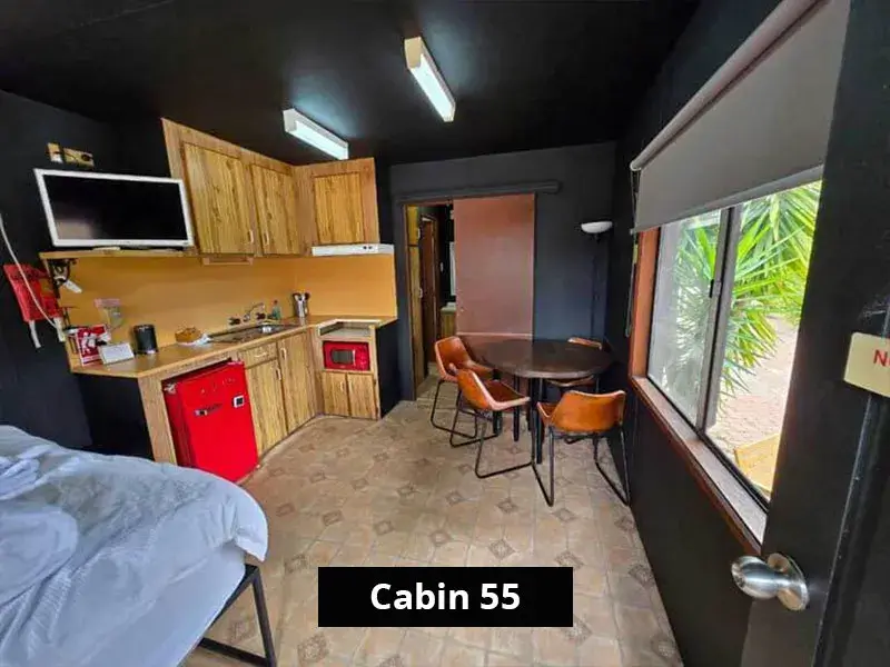 Dining and kitchen area inside Cabin 55, showing timber-look cabinetry, a red microwave and mini fridge, round dining table with four chairs, and large window with garden views.