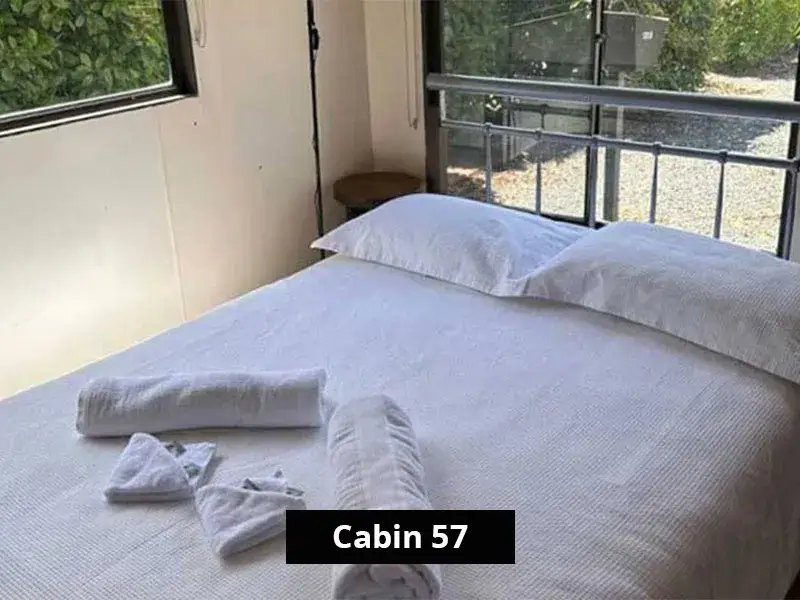 Interior bedroom of Cabin 57 at Stawell C&C Park, featuring a queen bed with white linen, pillows, and neatly folded towels, surrounded by large windows with views of greenery and the park's gravel pathway.