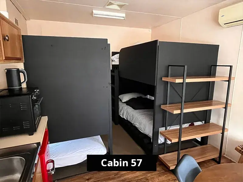 Sleeping area in Cabin 57 at Grampians Gate caravan park, showing two sets of black bunk beds, a small kitchenette with microwave and kettle, wood flooring, and wall-mounted air conditioning.