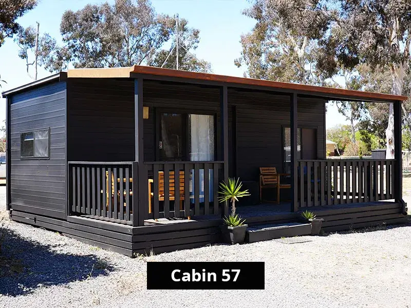 Exterior of Cabin 57 at Stawell C&C Park Grampians Gate, a dark timber cabin with a front deck, railing, seating, potted plants, and surrounded by trees and gravel pathways.