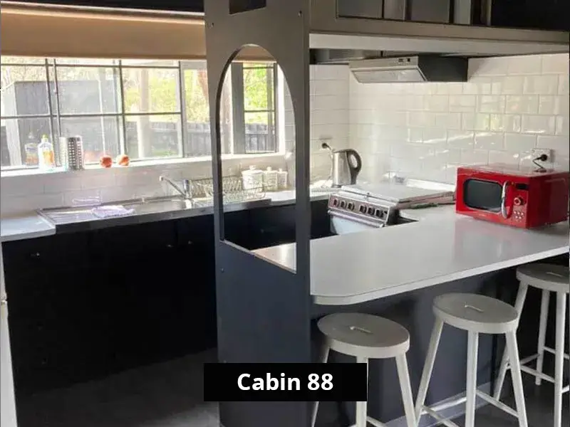 Full kitchen inside Cabin 88 featuring a modern cooktop, oven, red microwave, white subway tile splashback, and breakfast bar with stools beneath a large window.
