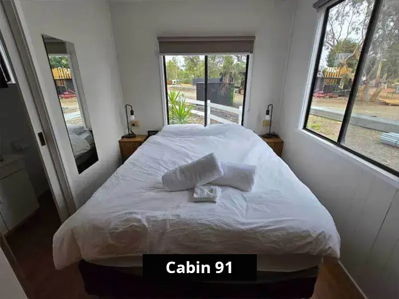 Bright queen bedroom inside Cabin 91 featuring large windows on two sides, white bedding with towels neatly arranged, two bedside tables with modern lighting, and a mirror next to a sliding door leading to the ensuite.