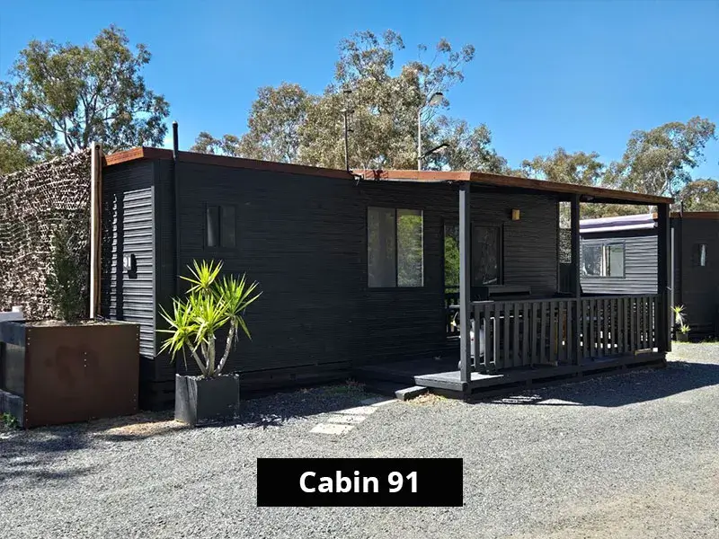 Exterior view of Cabin 91 at Grampians Gate caravan park in Stawell, showcasing a sleek black cabin with a covered front deck, outdoor seating, and surrounding trees in a peaceful park setting.