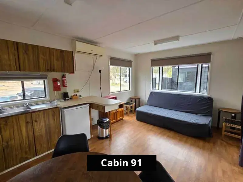 Open-plan interior of Cabin 91 with a compact kitchen, dining table, air conditioner, multiple windows, and a sofa bed in the lounge area, ideal for flexible short or long-term stays.