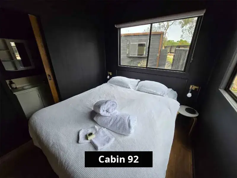 Queen bedroom in Cabin 92 with dark walls, two large windows providing natural light, and a neatly presented bed with fresh white linens and towels, plus bedside tables and pendant lights.