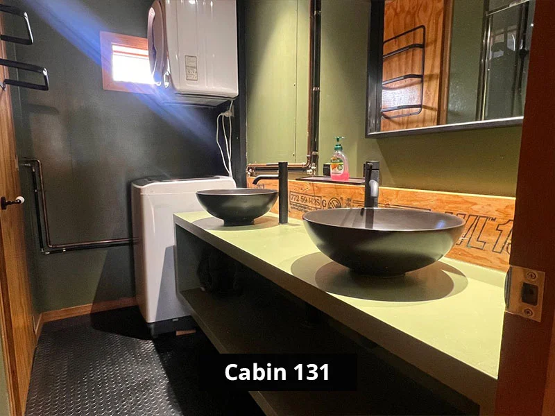 Cabin 131 bathroom with dual black vessel sinks, matte black faucets, and green countertop; features a compact washing machine and wall-mounted dryer under a small window, with industrial-style exposed piping and warm wood accents.