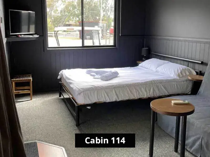 Bedroom in Cabin 114 featuring a queen bed with white linens, folded towels, large window with outdoor view, wall-mounted TV, and modern dark-toned walls.