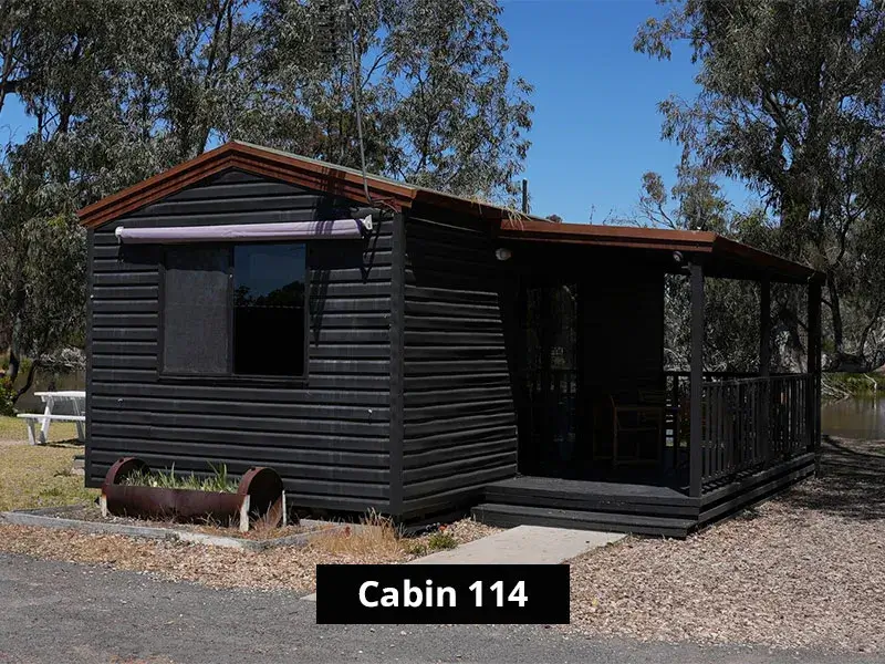 Exterior view of Cabin 114, a black wood-paneled vacation cabin with a covered porch, surrounded by trees and located near a water body.