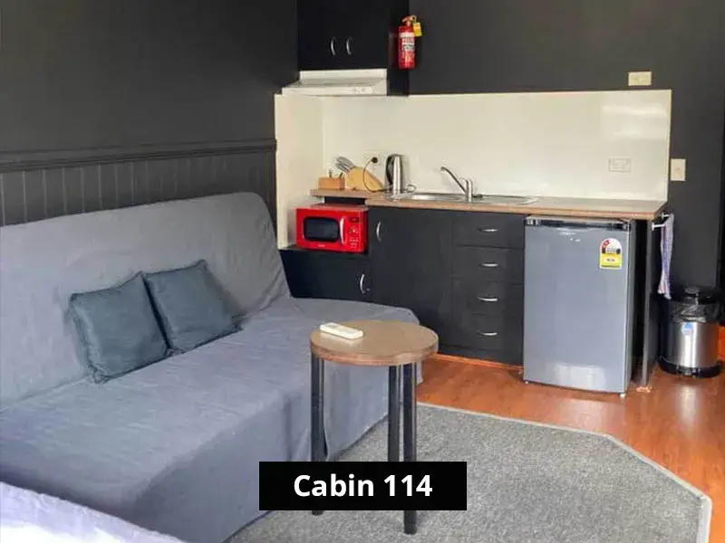 Compact kitchenette and lounge area in Cabin 114, featuring a grey sofa with cushions, mini fridge, red microwave, sink, countertop appliances, and black cabinetry.