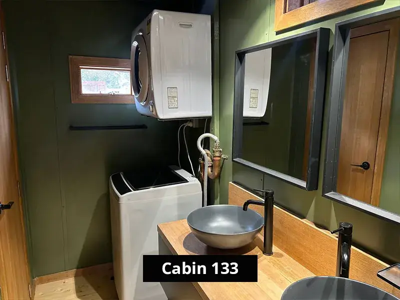 Modern rustic bathroom in Cabin 133 featuring a stacked washer and dryer unit, vessel sinks with matte black faucets, dual mirrors, and natural wood accents against olive green walls, blending functionality with stylish cabin decor.