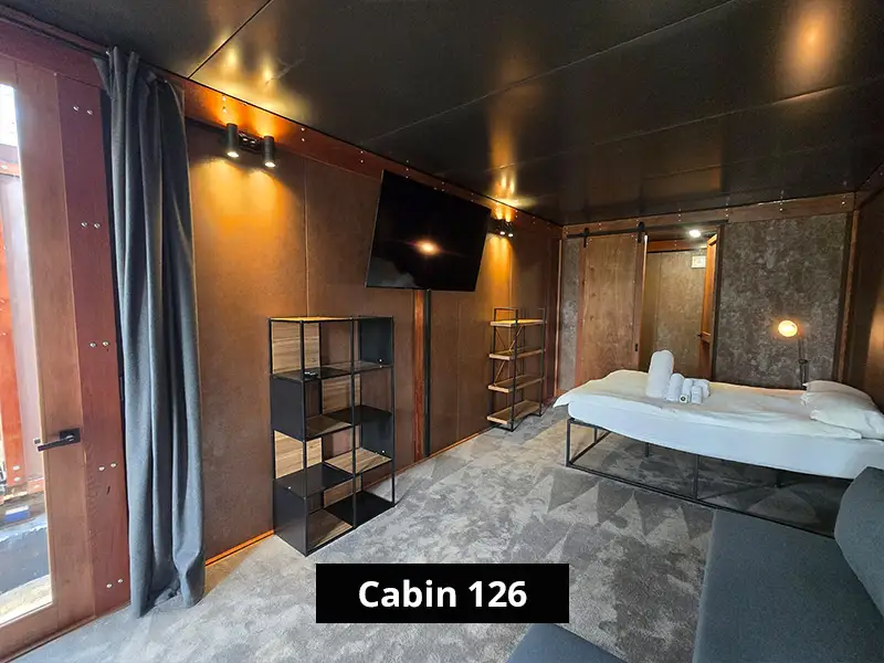 Minimalist cabin bedroom with double bed, wall-mounted TV, shelving units, and warm wood-paneled interior design – Cabin 126