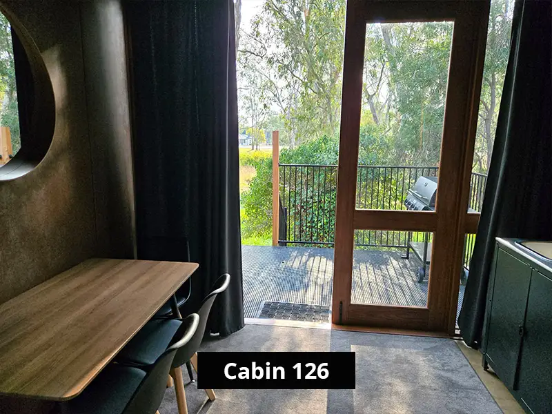 Small dining area inside cabin with table and chairs overlooking outdoor deck and greenery through glass doors – Cabin 126