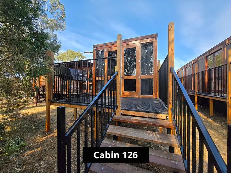 Modern wooden cabin with elevated deck, black railings, and stairs, surrounded by trees in a nature resort setting – Cabin 126 exterior