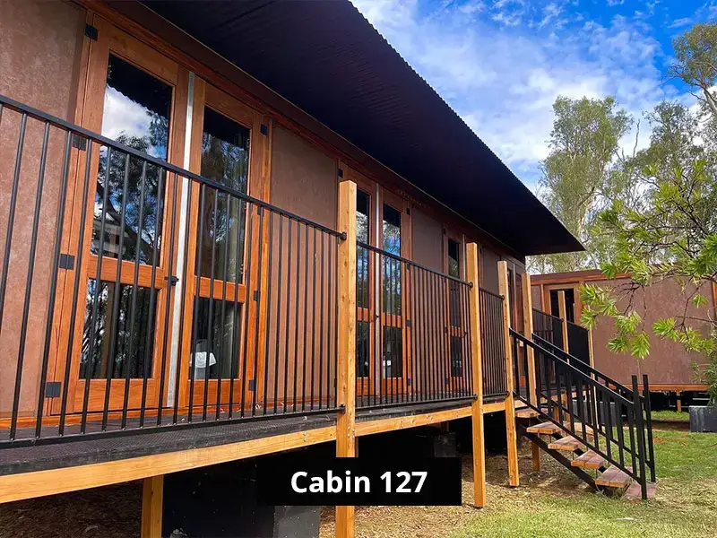 Modern wooden cabin exterior with elevated deck, black metal railings, large glass doors, and surrounding green trees under a blue sky.