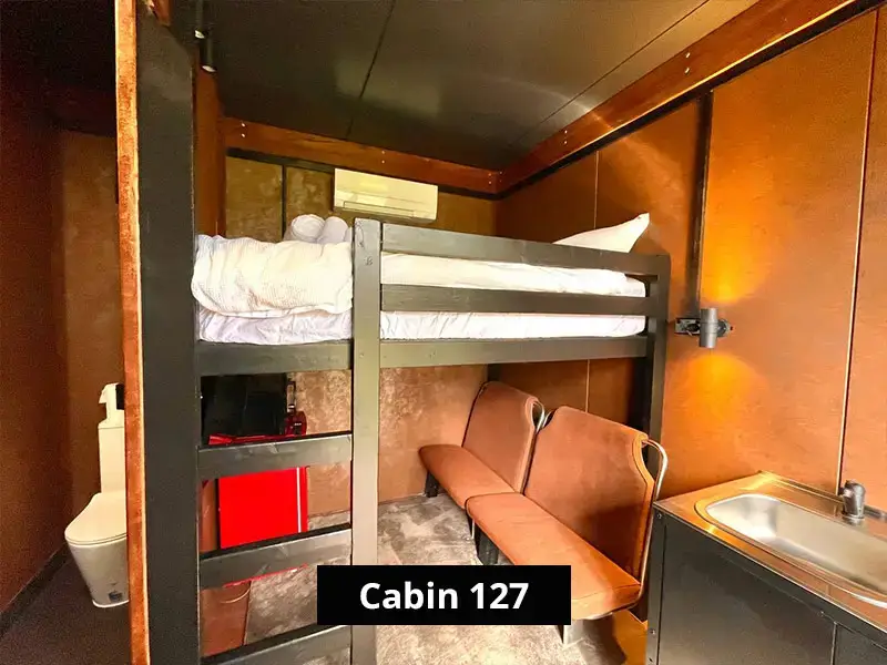 Compact cabin interior featuring bunk beds, small seating area, kitchenette with sink, and warm wooden walls for minimalist accommodation.