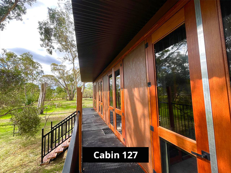 Cabin deck walkway with wooden posts, black railing, outdoor grill, and scenic grassy landscape with tall trees.