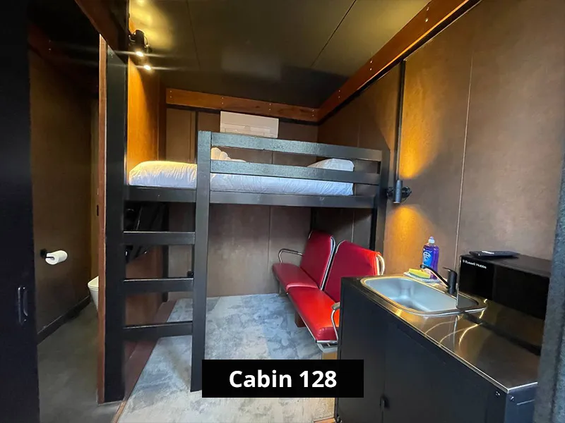 Small cabin interior with bunk beds, red seating bench, compact kitchenette with sink and microwave, and efficient space-saving design.