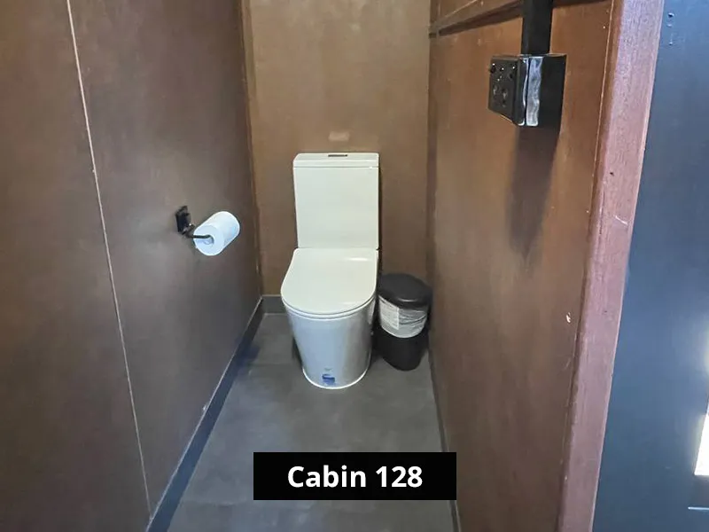 Compact cabin bathroom with modern toilet, toilet paper holder, small trash bin, and minimalist wood-paneled interior.