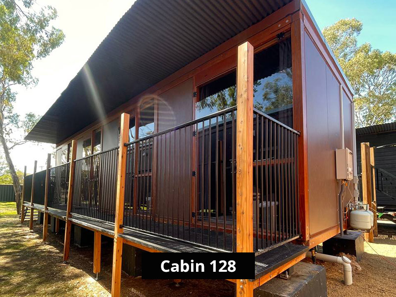 Modern prefab cabin exterior with elevated deck, black railings, large glass doors, and natural surroundings in a wooded setting.
