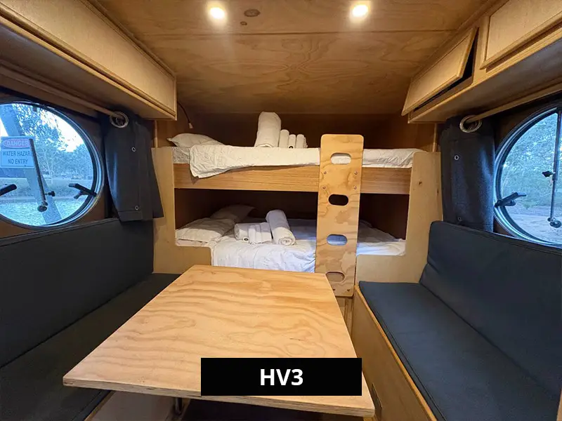 Interior view of HV3 with a small kitchenette, drawers, fridge, bunk beds to the left, and a fold-down dining table beside a round window with natural light.