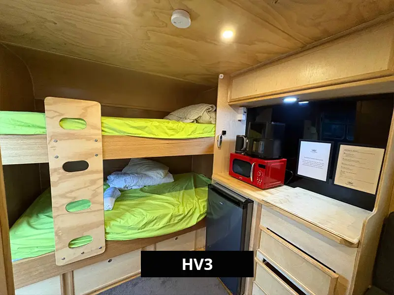 Interior of HV3 showing a double bunk bed setup with timber finish, white bedding, and a compact kitchenette area with mini fridge and storage cabinetry.