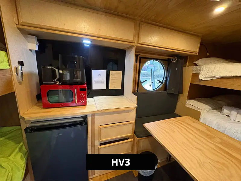 Compact living space in HV3 featuring bunk beds, a timber dining table with bench seating, and a circular window overlooking the lakeside setting.