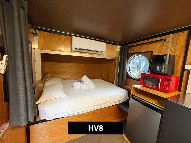 Interior of HV8 tiny home cabin with queen bed, timber walls, compact kitchen with sink, microwave, and round window views.