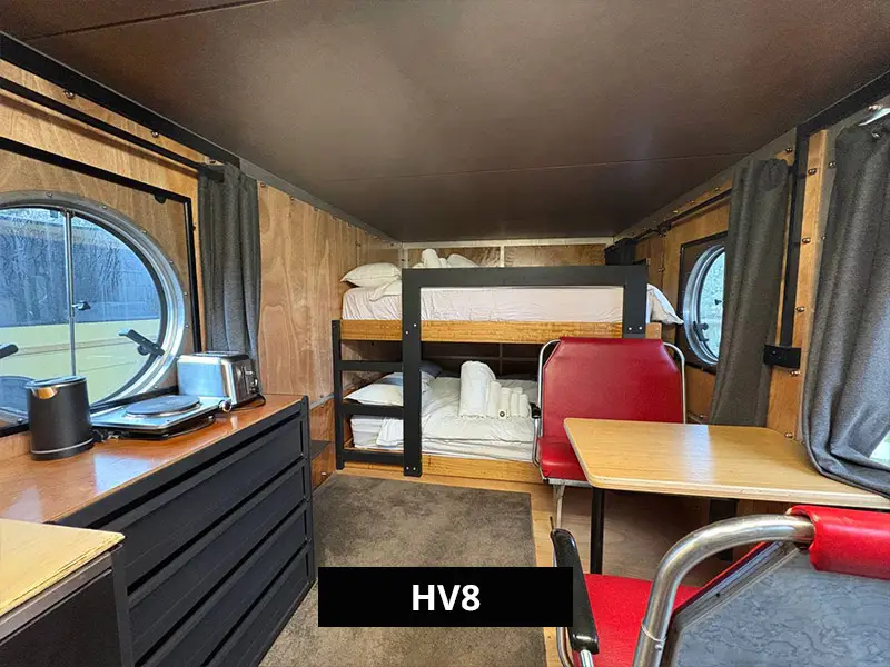 HV8 cabin interior featuring bunk beds, red retro-style dining booth, kitchenette, and round windows with natural light.